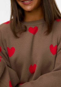 Pull en tricot marron avec des motifs de cœurs rouges. Le tissu a une texture douce, avec une coupe décontractée et des manches larges.