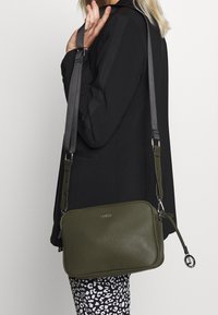 Oliven grøn læder crossbody taske med rektangulær form, justerbar grå rem, sikker lynlås og diskret branding på forsiden.