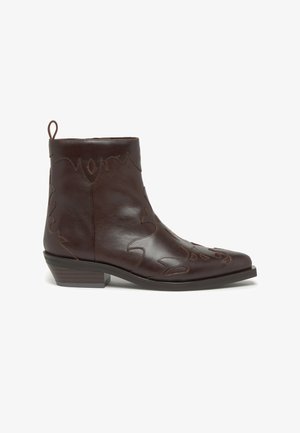 Bota de tobillo de cuero marrón con punta puntiaguda, costuras decorativas, tacón bajo apilado y lengüeta en la parte trasera. Textura lisa y diseño intrincado.