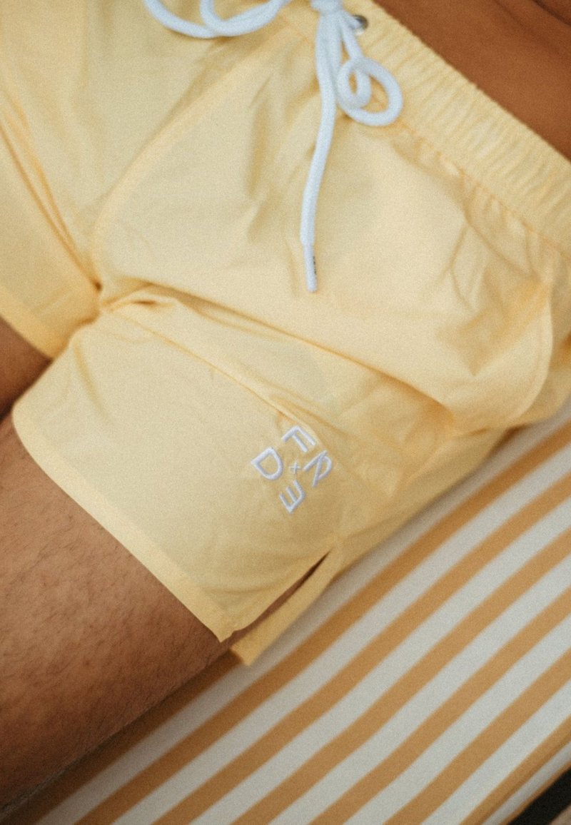 Pantaloni da bagno gialli con coulisse bianca e dettaglio del logo ricamato. Leggera lucentezza e texture liscia. Sfondo a righe in colori tenui.