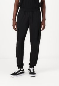 Sorte cargo-bukser med elastisk liv og sidelommer; tapered ben-design; kombinert med sorte sneakers med hvit såle og detaljer.