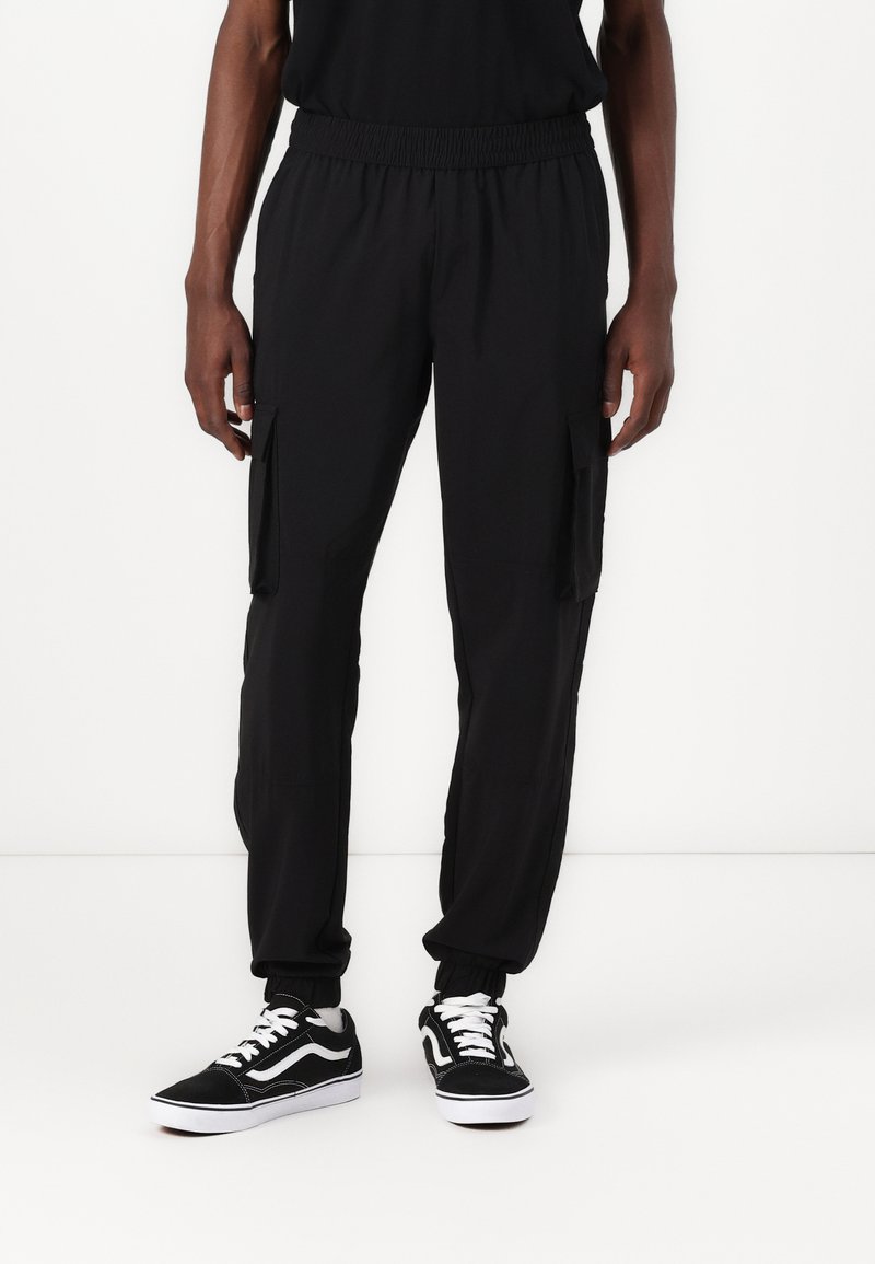 Sorte cargo-bukser med elastisk liv og sidelommer; tapered ben-design; kombinert med sorte sneakers med hvit såle og detaljer.