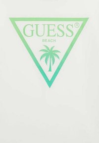 Camiseta blanca con un logo en forma de triángulo en degradado verde que incluye el texto "GUESS" y "BEACH", además de un gráfico de una palmera en el centro.