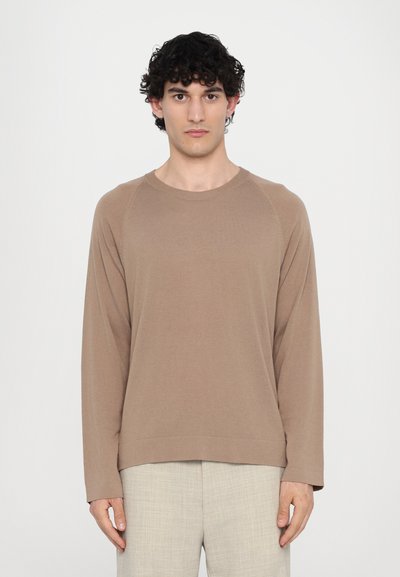 Sudadera marrón de manga larga con cuello redondo y mangas raglán, hecha de una tela suave. Combinada con pantalones de textura clara.