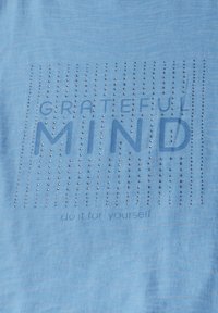T-shirt bleu clair avec "GRATEFUL MIND" et "do it for yourself" imprimés en texte foncé, agrémenté de clous métalliques en motif de grille.