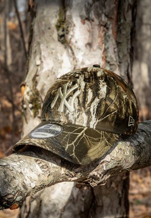 Camouflage baseball cap with a curved brim, featuring a raised light-colored New York logo, made of fabric with tree bark patterns.