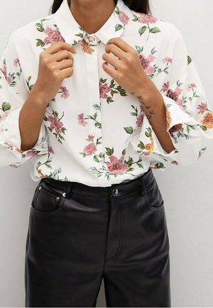 Femme ajustant le col d'une chemise blanche à manches longues avec imprimé floral rose, associée à un pantalon en cuir noir taille haute.