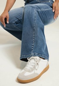 Denim flared jeans with silver studs along the seams, paired with white and gray sneakers featuring a gum sole. Textured fabric visible.