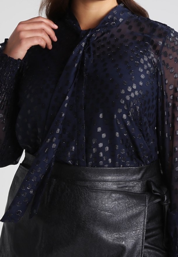 Marineblå gennemsigtig bluse med metallisk prikket mønster, lange ærmer og sløjfedetalje ved halsen, kombineret med en sort nederdel i imiteret læder.