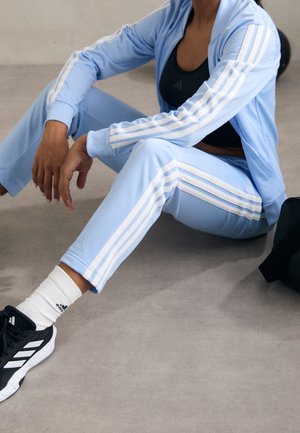 Person sidder på gulvet iført lyseblåt Adidas træningssæt, sort sports-BH, hvide sokker og sorte Adidas sneakers.