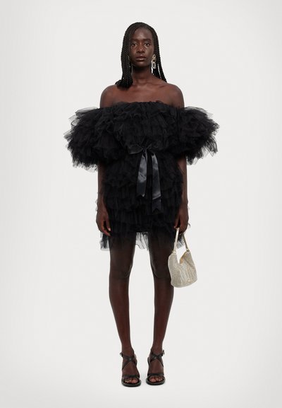 Zwarte tule jurk met off-the-shoulder ontwerp, gelaagde ruffles, satijnen riem en een zilveren net-handtas. Model draagt zwarte sandalen met bandjes.