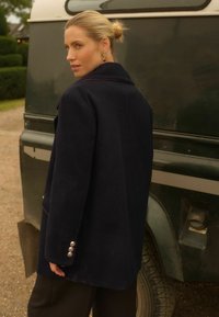 Námornícky vlněný kabát s přizpůsobeným designem, má klopový límec, zapínacie rukávy a kontrastní lemování. Pozadie obsahuje zelené vozidlo.