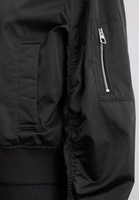 Black bomber jacket sleeve with ribbed cuff, side pocket, and small zippered pocket on upper arm against a neutral background.