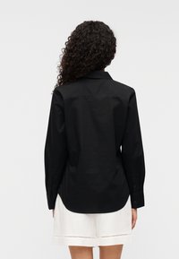 Μαύρο πουκάμισο με μακριά μανίκια από λείο ύφασμα, με κλασικό γιακά και μανσέτες με κουμπιά, εμφανίζεται από πίσω.