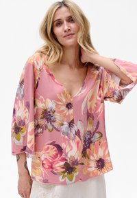 Kwiatowa bluzka z lekkiego różowego materiału, z rękawami dzwonkowymi i głębokim dekoltem w kształcie V. Wzory obejmują duże, kolorowe kwiaty w różnych odcieniach.