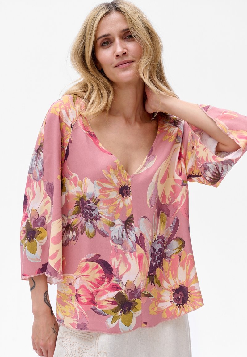 Kwiatowa bluzka z lekkiego różowego materiału, z rękawami dzwonkowymi i głębokim dekoltem w kształcie V. Wzory obejmują duże, kolorowe kwiaty w różnych odcieniach.