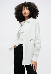 Kobieta o kręconych włosach, ubrana w białą koszulę zapinaną na guziki na białą bluzkę, czarne spodnie, stoi z jedną ręką w kieszeni na jednolitym tle.