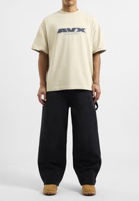 Camiseta de algodón beige con un logo azul de AVX, combinada con pantalones de mezclilla oscuros de corte holgado y calzado en tono marrón claro, completando un atuendo casual.