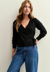 Svart omslagströja med långa ärmar, texturerad tyg och sidofäste, i kombination med ljusblå vida jeans.