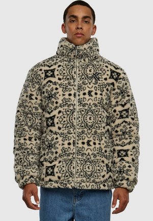 Chaqueta de forro polar en color crema con patrones intrincados en negro, cuello alto y cierre frontal con cremallera. Tejido texturizado con un aspecto acogedor y cálido.