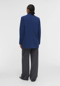 Blauwe blazer met schoudervullingen, lange mouwen en een rechte snit, gecombineerd met loszittende grijze broeken van een gladde stof.