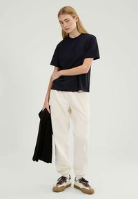 Marineblaues T-Shirt mit kurzen Ärmeln aus weicher Baumwolle, kombiniert mit cremefarbenen Jogginghosen. Das Model hält eine schwarze Jacke und vervollständigt das Outfit mit braunen Sneakern.