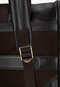 Hnědá kožená taška s texturovaným látkovým panelem, vybavená kovovou sponou a nastavitelným popruhem. Tmavé stehy zvýrazňují design.