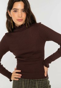 Jersey de punto acanalado marrón con cuello alto y mangas largas, con bordes con volantes y cintura ajustada. Combinado con pantalones de cuadros a cuadros.