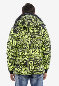 Schwarze Puffjacke mit großen neongrünen Logos und Textmustern. Mit Kapuze und gerippten Bündchen. Getragen über blauen Jeans.