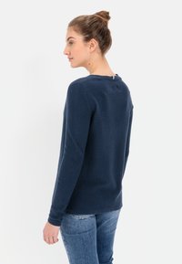 Marineblauwe geribde trui met lange mouwen en een ronde hals. Heeft een klein detail aan de achterkant bij de nek. Gecombineerd met blauwe spijkerbroeken.