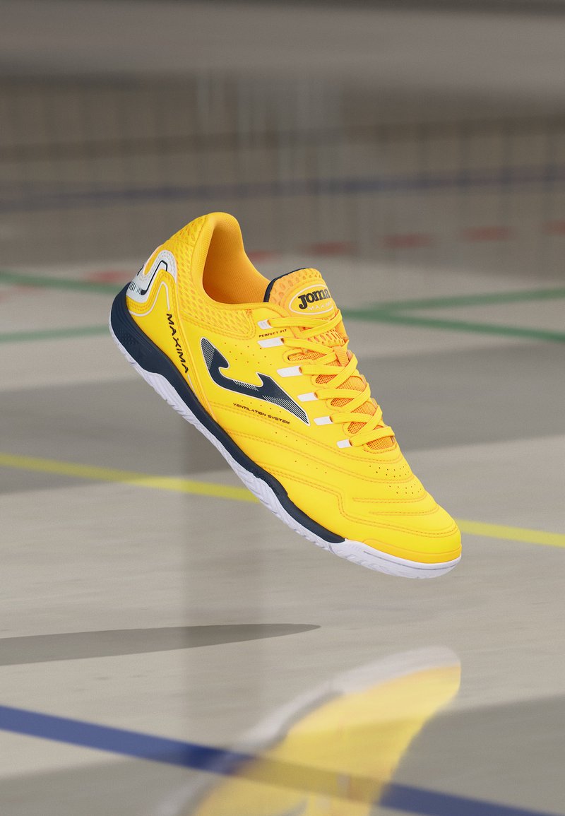 Zapatilla deportiva de interior de color amarillo brillante con suela negra y blanca, logo negro y texto "MAXIMA", suspendida sobre un suelo de gimnasio con líneas de colores.