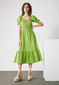 Grünes Baumwollkleid mit Puffärmeln, gerafftem Saum und elastischem Taillenbund. Getragen mit beigen Keilsandaletten mit Knöchelbindungen.