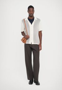 Man wearing short-sleeve buttoned shirt with vertical knit panels, dark wide-leg trousers, black shoes, and carrying tan crossbody bag.