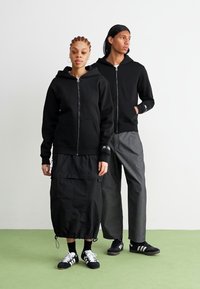 Sorte zip-up hættetrøjer med frontlommer, båret af to personer. En i en lagdelt sort nederdel, den anden i grå bukser og sneakers.
