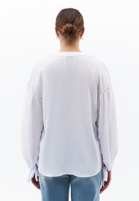 Blusa bianca fluida con maniche lunghe a sbuffo, orlo arrotondato e dettagli con lacci ai polsini. Tessuto leggero con una finitura testurizzata.