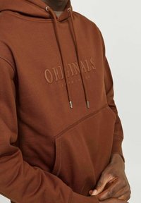 Sudadera marrón con un acabado texturizado, que cuenta con un bolsillo canguro, cordón ajustable y un logo en relieve "ORIGINALS" en el pecho.