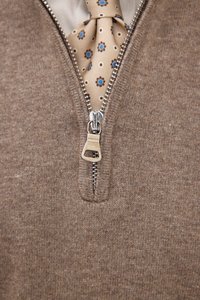 Bruine zip-up trui met een zilveren rits en een crèmekleurige kraag. Een crèmekleurige stropdas met blauwe en bruine bloemenpatronen is zichtbaar eronder.