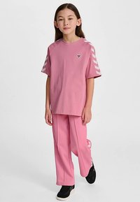 Rosa T-shirt med en logotyp på bröstet och matchande rosa sweatpants. Fiskbensmönster på ärmarna och sidan av byxorna. Svarta sneakers.