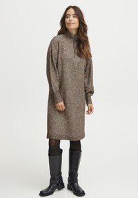 Robe en tricot marron avec un col montant et des manches longues, présentant des poignets et un ourlet côtelés. Portée avec des bottes noires jusqu'aux genoux.