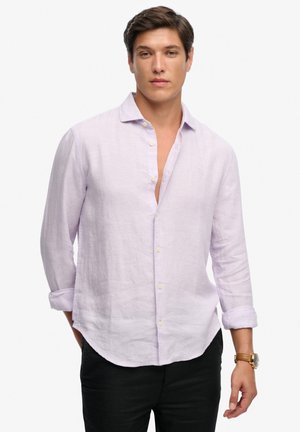 Mand iført lyse lilla langærmet hørskjorte med opkrogede ærmekanter, sorte bukser og armbåndsur på venstre hånd, stående mod en hvid baggrund.