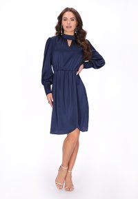 Granatowa satynowa sukienka z długimi bufkami, dekoltem w kształcie łezki i marszczoną talią. Posiada zwiewną spódnicę z lekko zakrzywionym dołem.
