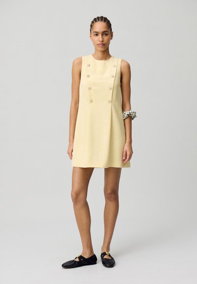 Abito giallo senza maniche con taglio dritto, caratterizzato da grandi bottoni beige. La modella indossa scarpe nere basse e ha un accessorio decorato sul braccio.