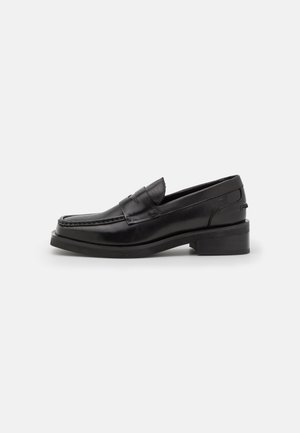 Mocasín de piel negra con puntera cuadrada redondeada, tacón bajo apilado y detalle clásico de tira para moneda en la parte superior lisa, mostrado sobre fondo blanco.