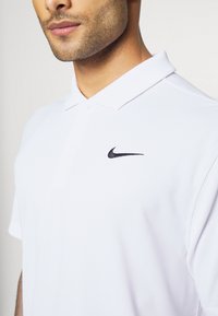 Baltos polo marškiniai, pagaminti iš lygaus audinio, su juoda Nike logo kairėje krūtinės pusėje ir klasikinėmis apykaklėmis bei dviem mygtukais.