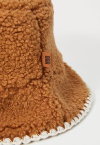 Brun fleece bucket-hat med teksturert overflate, kremfarget søm langs brettet, og en liten tan UGG-merkelapp festet.