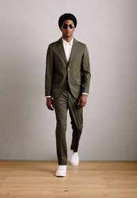Bărbat cu coafură afro poartă un costum verde măsliniu, cămașă albă, adidași albi și ochelari de soare, mergând pe un podea din lemn, în fața unui perete simplu.