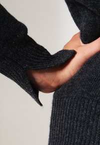 Donkere ribgebreide trui-arm met een textuur en zachte uitstraling, voorzien van een uitsnede bij de pols die een hand onthult.