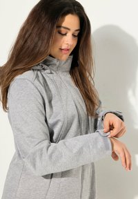 Chaqueta gris con cremallera y cuello alto, mangas largas y puños ajustables. Hecha de un tejido suave y texturizado con detalles de costura.