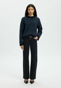 Sieviete ar gariem viļņotiem matiem, kam uzvilkts tumši zils teksturēts džemperis, melni augsto jostasvietu džinsi, melna josta un norādīti melni apavi, stāv.