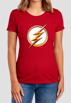 Woman wearing a red t-shirt with a gold lightning bolt symbol inside a white circle on the chest, paired with blue jeans.
